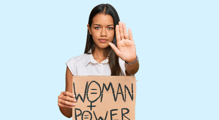 Canvas Print - Beautiful hispanic woman holding woman power banner with open hand doing stop sign with serious and confident expression, defense gesture