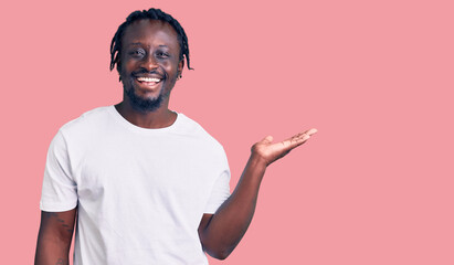 Poster - Young african american man with braids wearing casual white tshirt smiling cheerful presenting and pointing with palm of hand looking at the camera.