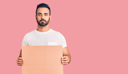 Poster - Young hispanic man holding banner cardboard thinking attitude and sober expression looking self confident