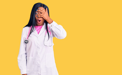 Poster - Young african american woman wearing doctor stethoscope peeking in shock covering face and eyes with hand, looking through fingers with embarrassed expression.