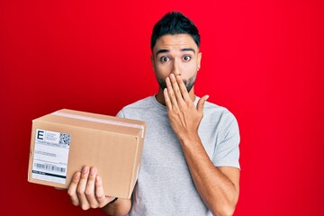 Wall Mural - young man with beard holding delivery package covering mouth with hand, shocked and afraid for mista