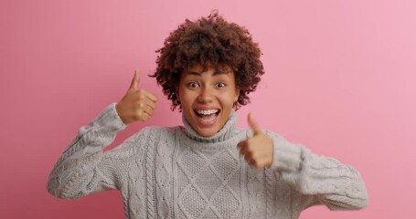 Wall Mural - Beautiful joyful dark skinned woman shows thumbs up with both hands rates something makes like gesture glad to achieve goals poses in casual jumper against rosy background. Only positive answer