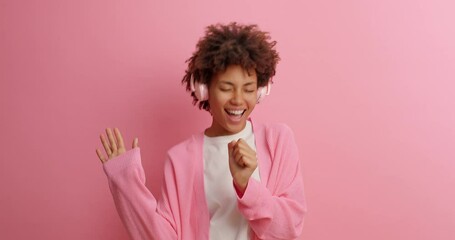 Wall Mural - Overjoyed dark skinned ethnic woman moves with rhythm of music wears headphones and sings favorite song dressed in casual clothing isolated over pink background. Slow motion. Carefree dance.