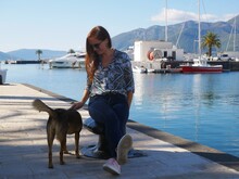 A Girl In Sunglasses, Dressed In Dark Jeans And A Green Shirt, Sits On The Dock And Strokes A Dog