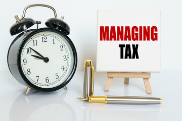 On the table there is a clock, a pen and a stand with a card on which the text is written - MANAGING TAX