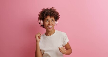 Wall Mural - Merry curly haired woman has upbeat mood dances carefree listens pleasant music in headphones moves with rhythm sings song keeps hand near mouth as microphone wears basic white t shirt poses indoor