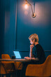 © qunica.com - Confident young businesswoman working on her laptop while sitting in a corner of a cafe with dark blue walls.