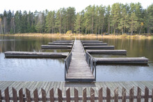 The Pier And Marina On Pontoons Reflecting In The Water, With A Walkway Between The Handrails Against The Backdrop Of A Pine Forest.