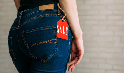 Wall Mural - People, sale, consumerism concept. Close up cropped photo of sexual woman's buttocks wearing blue casual denim pants on gray brick background in the mall. Red tag black Friday on jeans.