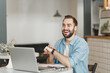 © ViDi Studio - Cheerful funny young man sitting alone at table in coffee shop cafe restaurant indoors working or studying on laptop pc computer using smart watch on hand. Freelance mobile office business concept.