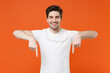 © ViDi Studio - Smiling cheerful young man 20s wearing basic casual empty blank white t-shirt standing pointing index fingers down on mock up copy space isolated on bright orange colour background, studio portrait.