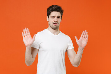 Wall Mural - Displeased shocked worried nervous young man 20s in basic casual white t-shirt standing rising hands up showing palms looking camera isolated on bright orange colour wall background, studio portrait.