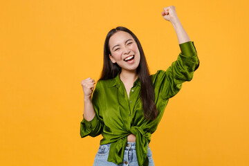 Wall Mural - Happy overjoyed funny young brunette asian woman 20s wearing basic green shirt standing clenching fists doing winner gesture looking camera isolated on bright yellow colour background studio portrait.