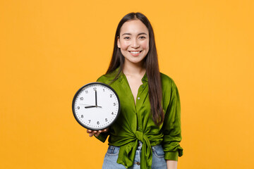 Smiling pretty attractive beautiful young brunette asian woman 20s wearing basic green shirt standing holding in hand clock looking camera isolated on bright yellow colour background, studio portrait.