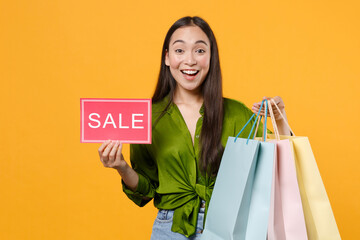 Wall Mural - Excited young brunette asian woman 20s in basic green shirt standing hold package bags with purchases after shopping sign with SALE title looking camera isolated on yellow background, studio portrait.