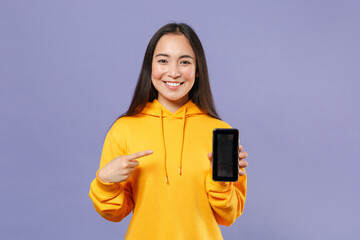 Wall Mural - Smiling young brunette asian woman 20s in basic yellow hoodie standing pointing index finger on mobile cell phone with blank empty screen isolated on pastel violet colour background, studio portrait.