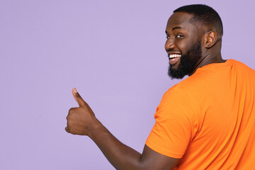 Back rear view of smiling young african american man 20s wearing basic casual orange blank empty t-shirt standing showing thumb up looking camera isolated on pastel violet background, studio portrait.