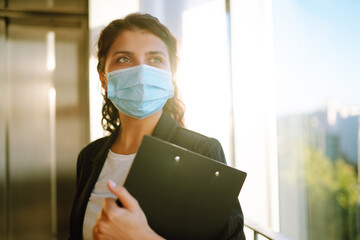 Young business woman wear face mask protect of coronavirus standing near elevator at modern office. Covid-19.