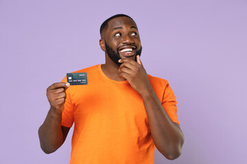 Wall Mural - Pensive young african american man wearing basic casual orange blank empty t-shirt standing hold credit bank card put hand prop up on chin isolated on pastel violet colour background studio portrait.