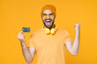 © ViDi Studio - Happy joyful young bearded man 20s in basic casual t-shirt headphones eyeglasses hat standing hold credit bank card doing winner gesture looking camera isolated on yellow background, studio portrait.