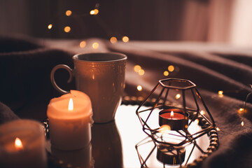 Cup of hot tea with burning candles on tray in bed over Christmas lights close up. Night time atmosphere at home.