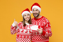 Smiling Young Santa Couple Friends Man Woman 20s In Red Sweater Christmas Hat Hold Gift Certificate Showing Thumb Up Isolated On Yellow Background. Happy New Year Celebration Merry Holiday Concept.