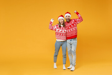 Full length happy young Santa couple friends man woman in sweater, Christmas hat doing winner gesture hugging isolated on yellow background studio portrait. Happy New Year celebration holiday concept.