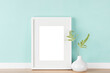 Elegant poster artwork mockup template for online shop with vertical picture frame with matte and vase with fern leaves in front of pastel green wall. Blank image area isolated with clipping path.