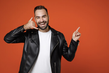 Wall Mural - Laughing young bearded man in basic white t-shirt black leather jacket doing phone gesture like says call me back pointing index finger aside up isolated on orange colour background studio portrait.