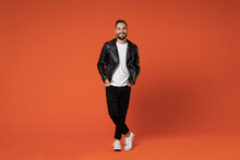 Full Length Of Smiling Cheerful Young Bearded Man 20s Wearing Basic White T-shirt Black Leather Jacket Standing Holding Hands In Pockets Looking Camera Isolated On Orange Background Studio Portrait.