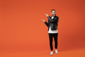 Full length of excited young bearded man in basic white t-shirt black leather jacket standing pointing index fingers aside on mock up copy space isolated on orange colour background studio portrait.