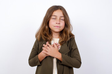 Wall Mural - Beautiful little girl standing against white backgr closes eyes and keeps hands on chest near heart, expresses sincere emotions, being kind hearted and honest. Body language and real feelings concept.