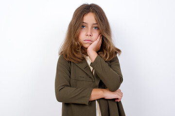 Wall Mural - Very bored Beautiful little girl standing against white background, holding hand on cheek while support it with another crossed hand, looking tired and sick.