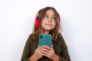 Wall Mural - Happy Beautiful little girl standing against white background, listening to music with earphones using mobile phone.