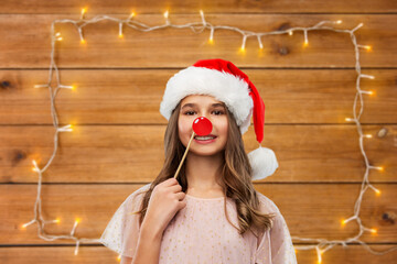 christmas party, holidays and photo booth concept - happy smiling teenage girl in santa helper hat with red nose accessory over garland lights on wooden background