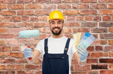 Poster - profession, construction and building concept - happy smiling male worker or builder in yellow helmet and overall with paint roller and color palettes over brick wall background