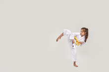 Full Length Shot Of Little Karate Girl In White Kimono With A Yellow Sash Exercising And Fighting, Doing Martial Arts, Standing Isolated Over White Background