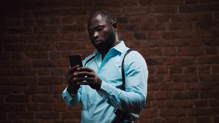 Confident african detective with holsters using smartphone isolated on red brick wall background