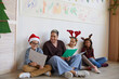 © Seventyfour - Full length portrait of female teacher sitting on floor with multi-ethnic group of kids holding pictures while enjoying art class on Christmas, copy space