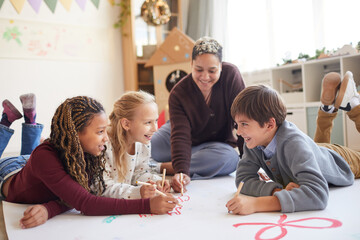 Full length portrait of smiling female teacher sitting on floor with multi-ethnic group of kids drawing pictures while enjoying art class, copy space