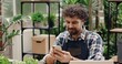 © VAKSMANV - Close up portrait of cheerful Caucasian male entrepreneur texting on smartphone while sitting in flower shop in apron. Happy man employee typing on cellphone at work. Floral concept
