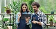 © VAKSMANV - Portrait of young Caucasian lovely couple working in own flower shop together. Joyful female and male entrepreneurs in aprons typing on tablet and talking in floral store. Family business concept