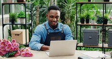 Joyful Young Man In Flower Shop Sitting At Workplace And Texting On Computer. Cheerful African American Male Florist Worker In Floral House Tapping And Typing On Laptop. Floristry Concept