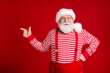 Wall Mural - Photo of retired old man grey beard direct finger empty space smile promoting winter outfit wear santa x-mas costume suspenders spectacles striped shirt cap isolated red color background