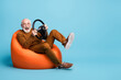 © deagreez - Portrait of his he nice attractive cheerful cheery glad funny bearded grey-haired man sitting in bag chair having fun holding in hands steering wheel isolated over blue pastel color background