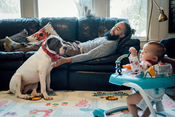 Man relaxing on couch petting dog while baby plays nearby