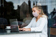 © Mint Images - Woman wearing face mask sitting alone at a cafe table with a laptop computer, working remotely.
