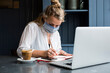 © Mint Images - Woman wearing face mask sitting in a cafe with a laptop working remotely.