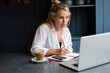 © Mint Images - Woman sitting alone at a cafe table with a laptop computer, writing in note book