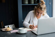 © Mint Images - Woman sitting alone at a cafe table with a laptop computer, writing in note book, working remotely.